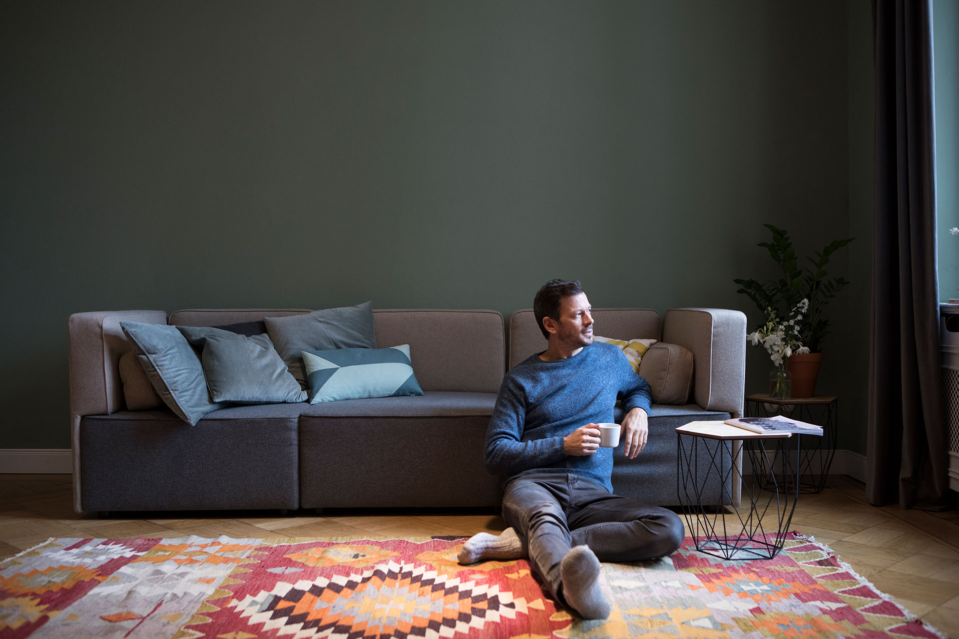 A relaxed man sits on the floor next to a couch, leaning back with a cup in hand, looking to the side. A cozy living room setting with cushions on the sofa.