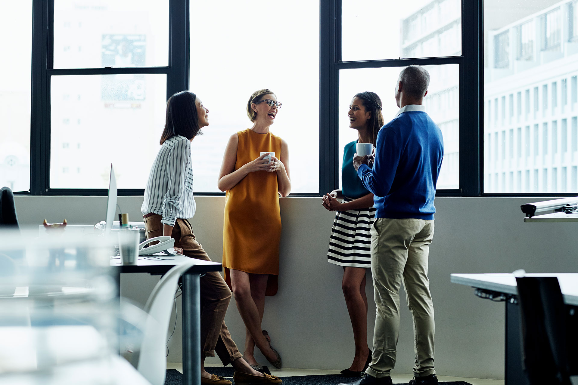Four business professionals are engaged in an informal meeting. They are conversing and smiling with two holding coffee cups, conveying a warm, collegial atmosphere.