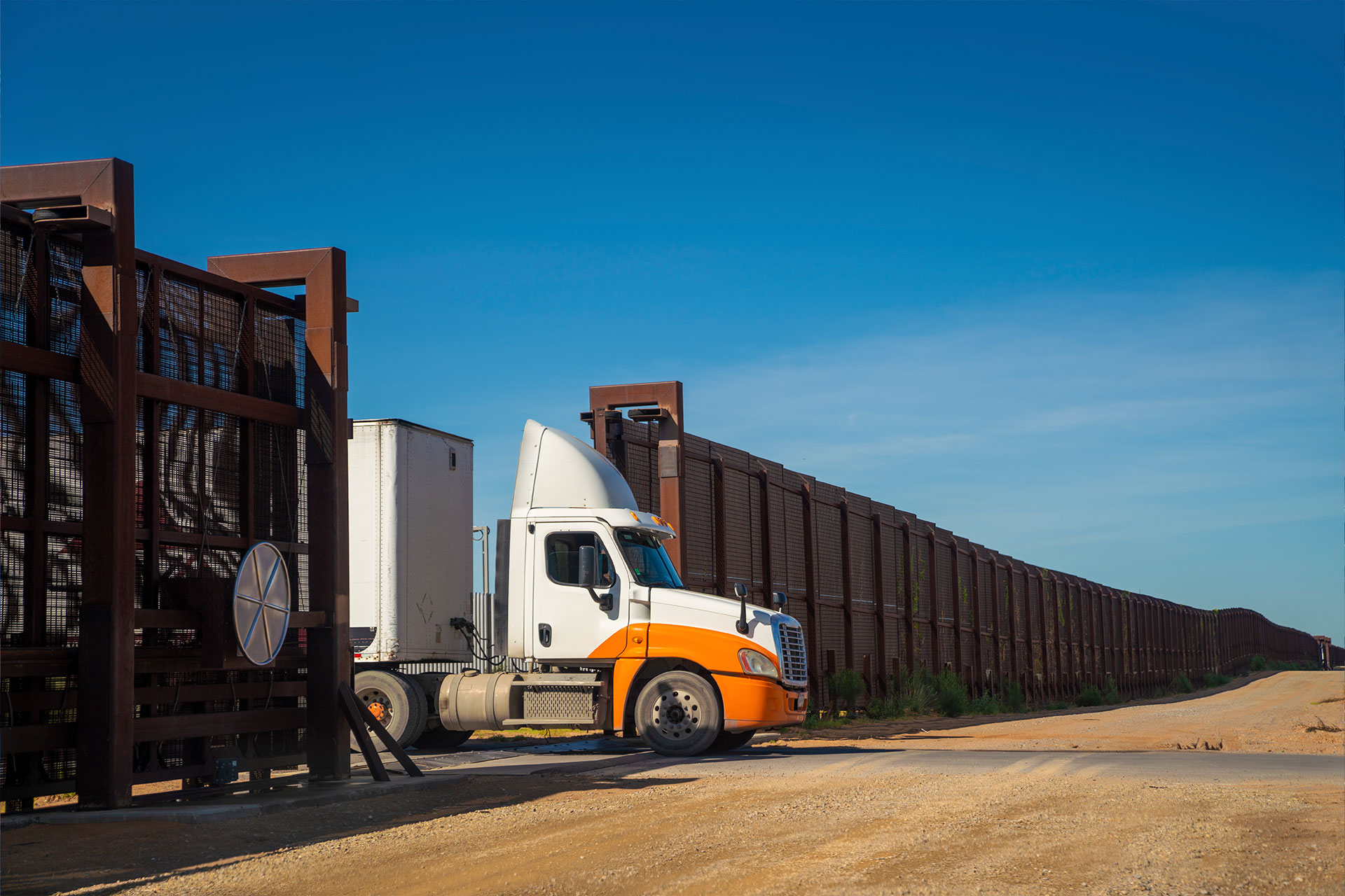 Photo of a truck driving through a metal gate with a long fence in the background.
